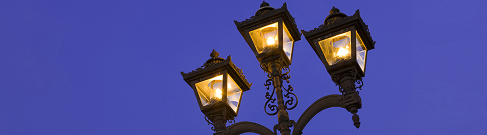 Three ornate gaslamp-style streetlights glowing against a clear evening sky.