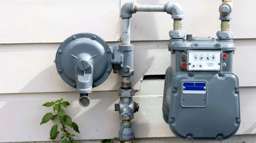 Gray residential gas meter with pipes attached to the exterior wall of a beige house.