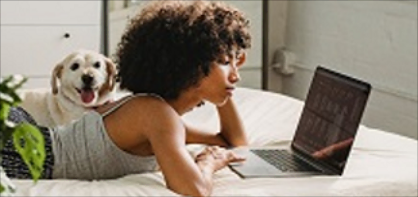 Woman lying on bed using a laptop with a happy dog beside her.