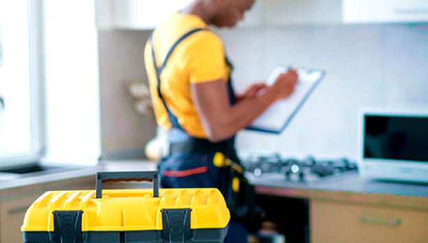 Person in yellow shirt and overalls writing on clipboard near kitchen counter with a yellow and black toolbox in the foreground.