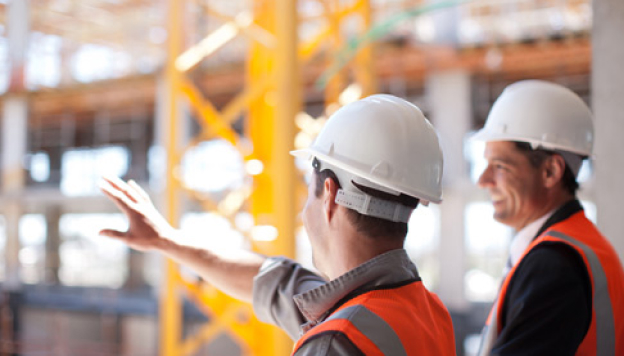 Two construction workers wearing white helmets and orange safety vests discussing plans at a construction site with scaffolding and yellow structural elements.
