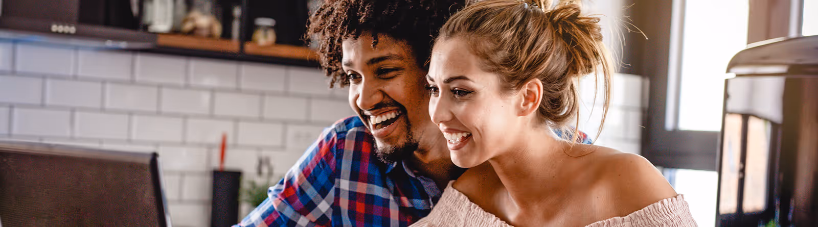 Smiling couple looking at a laptop screen in a kitchen with white subway tile backsplash.