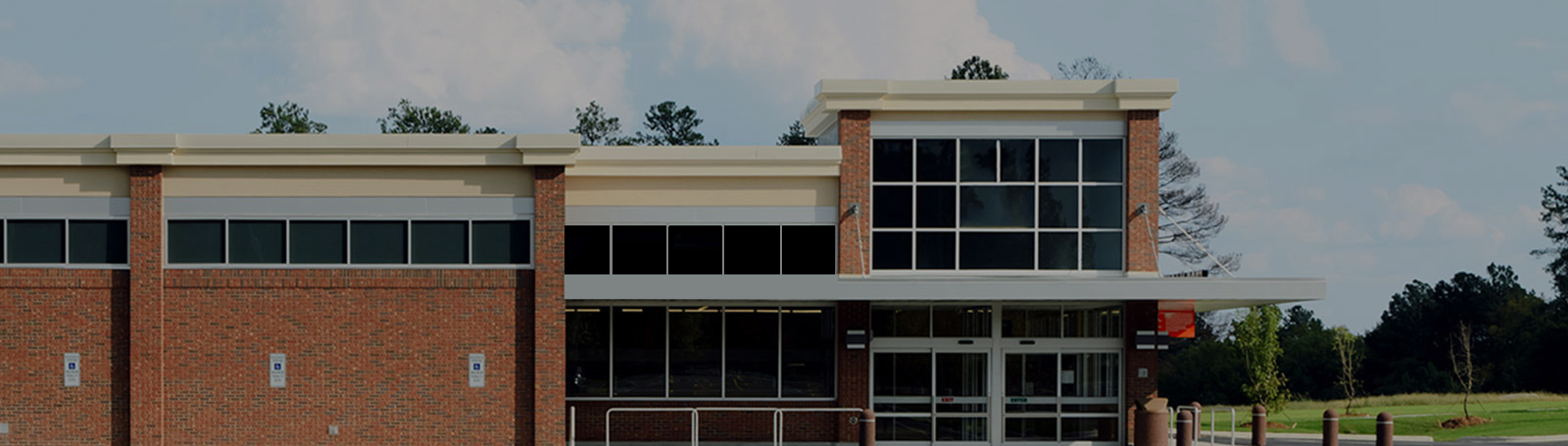Modern brick building with large windows and glass entrance doors, surrounded by greenery under a partly cloudy sky.