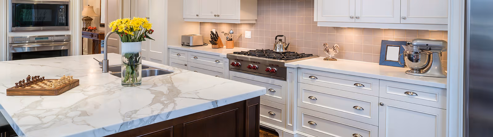 Modern kitchen with marble island countertop, vase of yellow flowers, chessboard, stainless steel appliances, and white cabinetry.