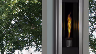 Modern fireplace with visible flames inside a sleek vertical metal frame, next to a window showing green tree leaves outside.