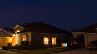Single-story house at night with warmly lit windows and porch light in a quiet neighborhood.