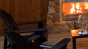 Two black wooden chairs on a wooden deck facing a stone fireplace with a fire burning inside.