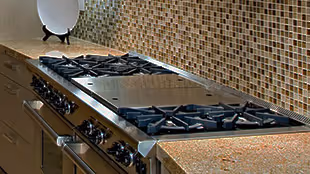 Stainless steel kitchen stovetop with multiple gas burners and a built-in griddle under a mosaic tile backsplash.