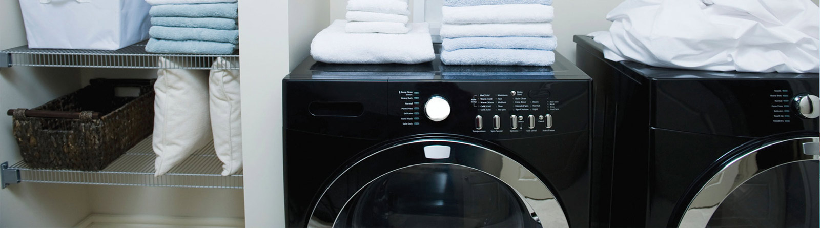 Two black front-loading washing machines with folded white and blue towels on top and shelving with pillows and baskets beside them.