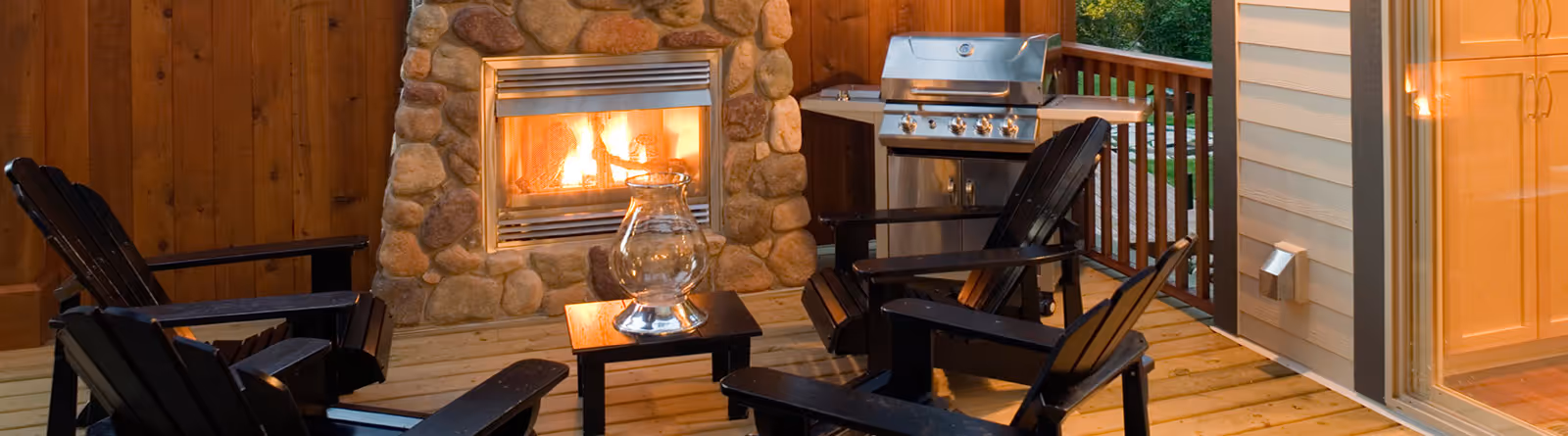 Cozy outdoor patio with lit stone fireplace, four black wooden chairs, small table with glass lantern, and stainless steel grill.