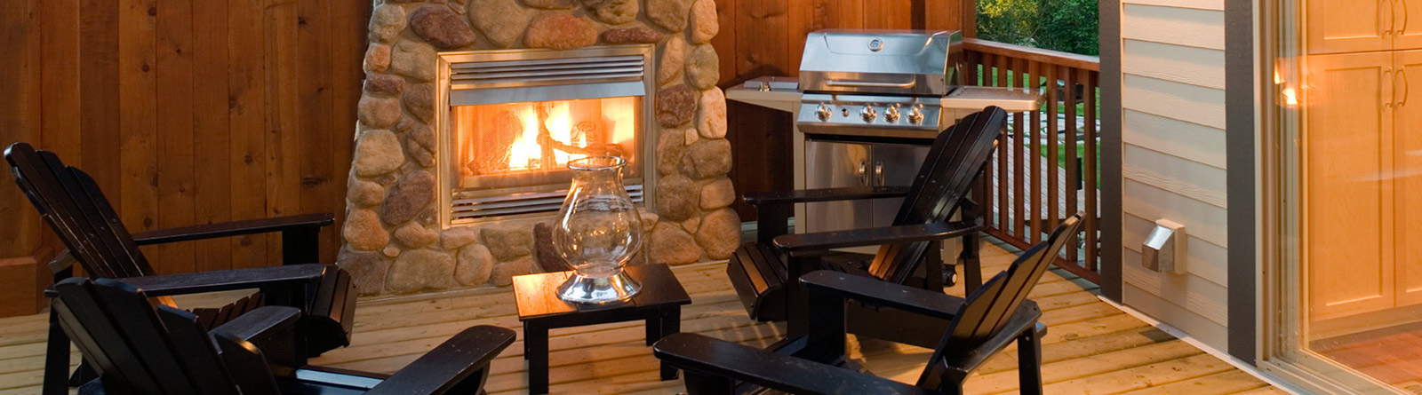 Cozy outdoor patio with lit stone fireplace, four black wooden chairs, small table with glass lantern, and stainless steel grill.