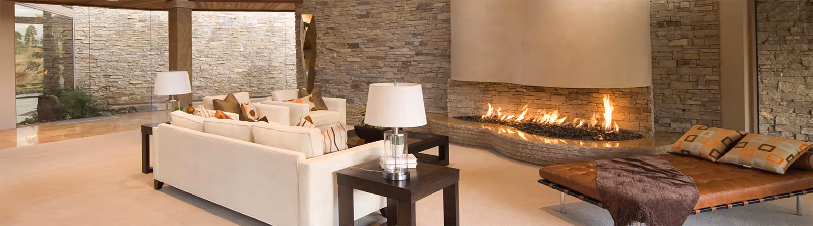 Modern living room with beige couches, a long fireplace with stone wall, and a leather daybed with pillows and a throw.