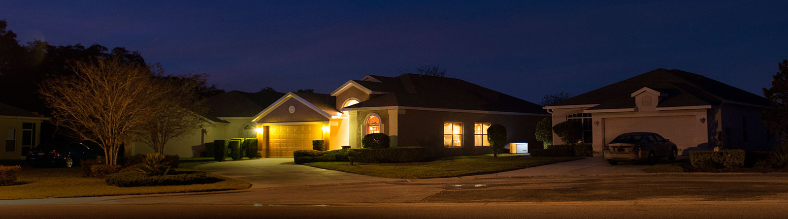 Suburban houses at dusk with illuminated garage and warm indoor lights visible through windows.