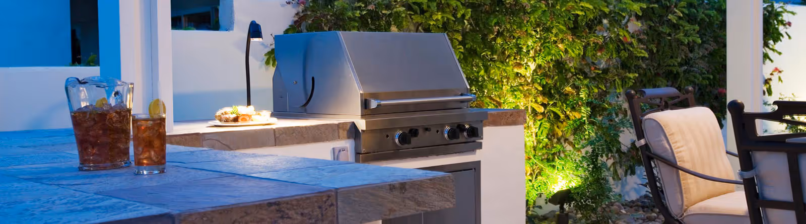 Outdoor kitchen with a built-in stainless steel grill, stone countertop, pitcher and glass of iced tea, and cushioned patio chairs near greenery illuminated by garden lights.