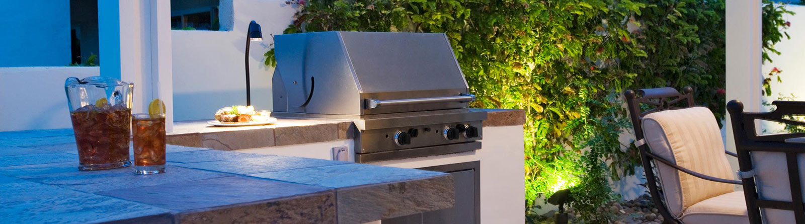 Outdoor kitchen with a built-in stainless steel grill, stone countertop, pitcher and glass of iced tea, and cushioned patio chairs near greenery illuminated by garden lights.
