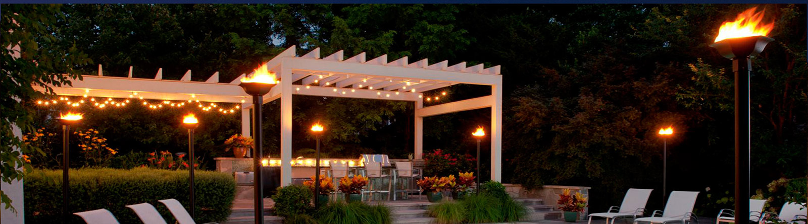 Outdoor patio with white pergola, string lights, flaming torches, and seating area surrounded by greenery at night.