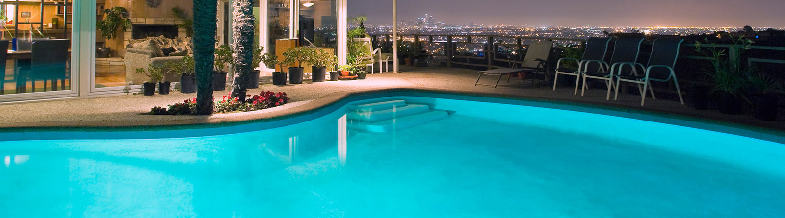 Nighttime view of a lit swimming pool next to a patio with plants and chairs, overlooking a city skyline with lights.