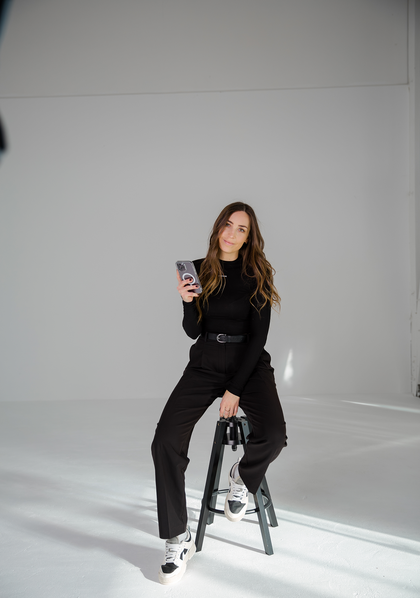Frau in schwarzem Oberteil und Hose sitzt auf einem Hocker und hält ein Smartphone in der Hand.