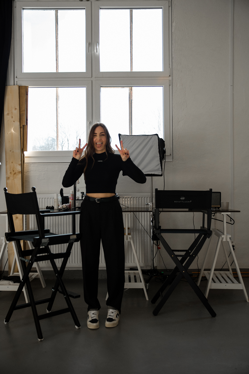 Frau in schwarzem Outfit macht Frieden-Zeichen mit beiden Händen vor zwei schwarzen Regiestühlen in einem hellen Raum mit großen Fenstern.