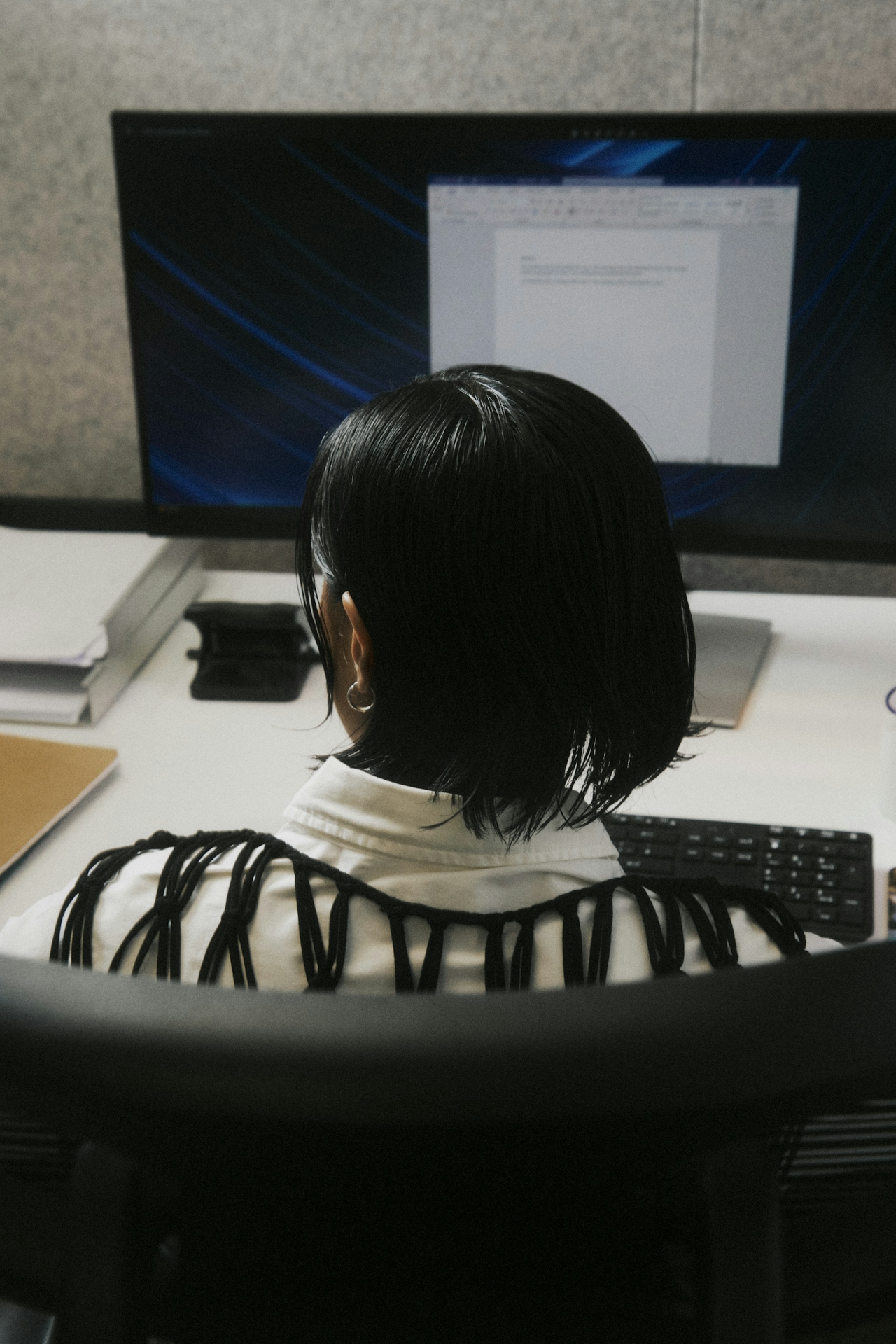 Person mit kurzen schwarzen Haaren sitzt vor einem Computerbildschirm mit einem geöffneten Dokument in einem Büro.