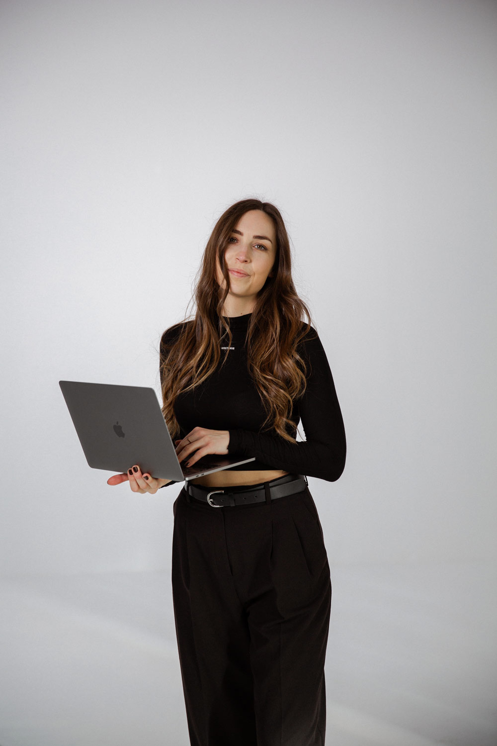 Junge Frau mit langen braunen Haaren trägt schwarze Kleidung und hält einen aufgeklappten Laptop.