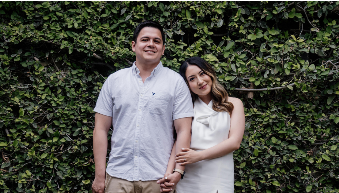 Pareja sonriente posando frente a una pared de hojas verdes en estilo clásico. Ambos visten ropa clara e informal.