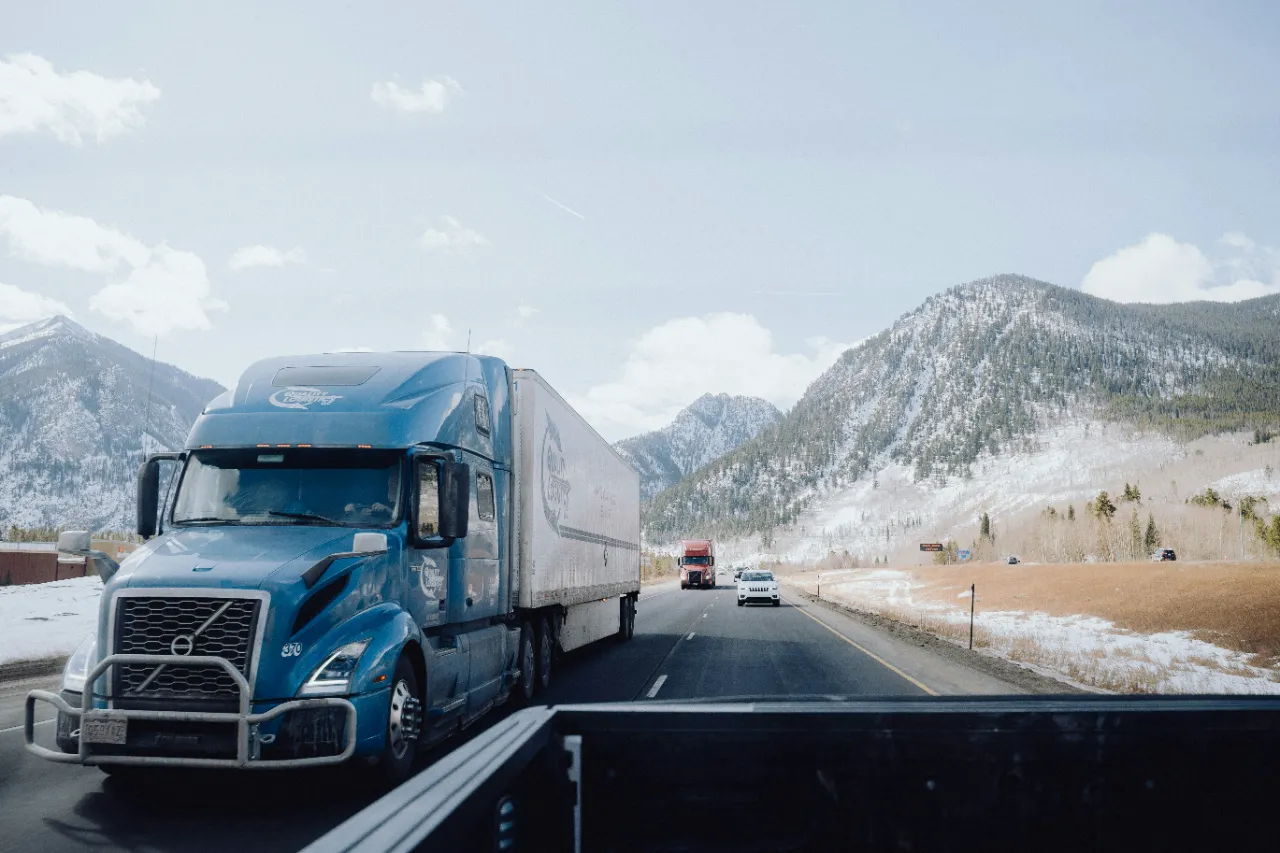 Blue semi-truck hauling a trailer along a snowy mountain highway, with surrounding peaks, winter landscape, and passing traffic under clear skies.