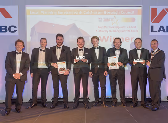 Seven men in black tuxedos standing on a stage holding awards, with a presentation slide behind them showing Colchester Borough Council recognition.