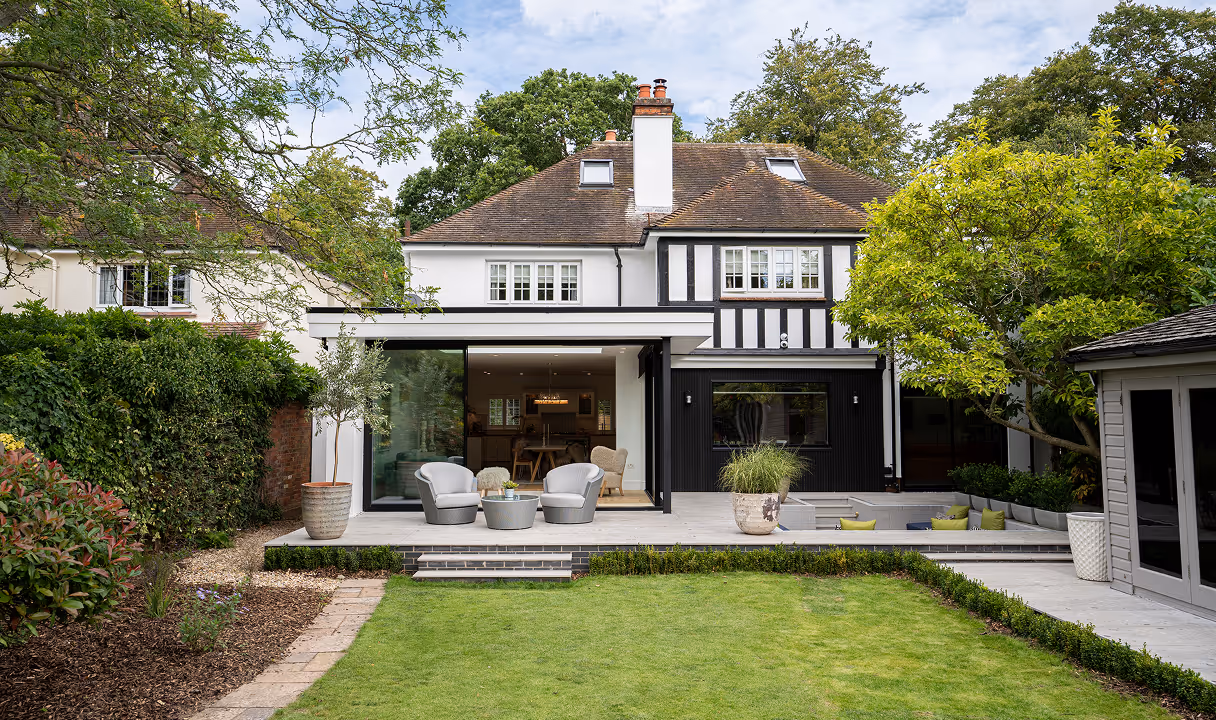 Modern two-story house with a spacious patio featuring outdoor chairs, potted plants, and a sunken seating area surrounded by greenery.