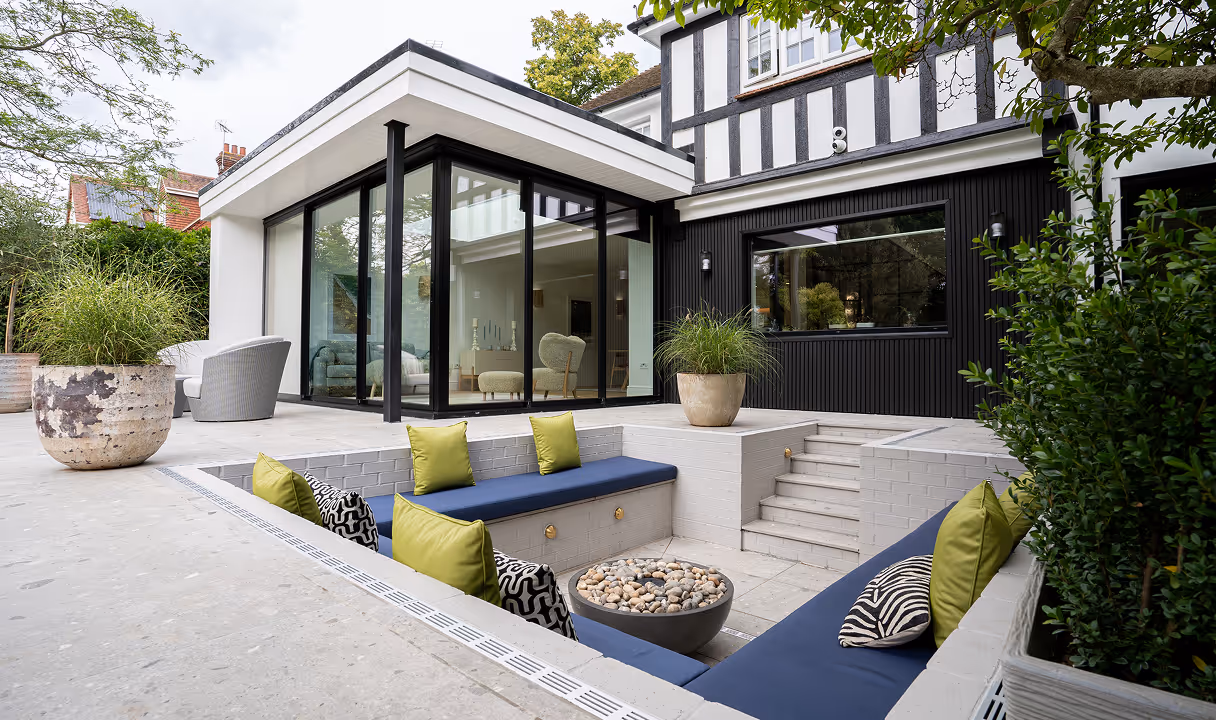 Modern outdoor sunken seating area with navy cushions and green pillows surrounding a round stone fire pit, adjacent to a house with large glass windows and black and white exterior.