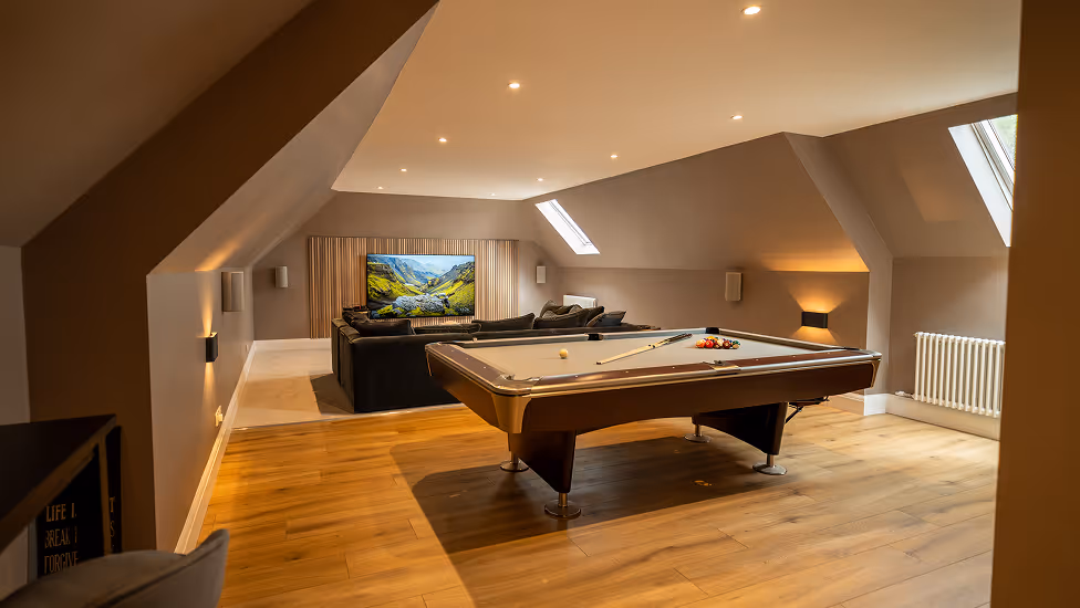 Spacious attic game room with a pool table, black sectional sofa, wall-mounted TV, and slanted ceiling windows.