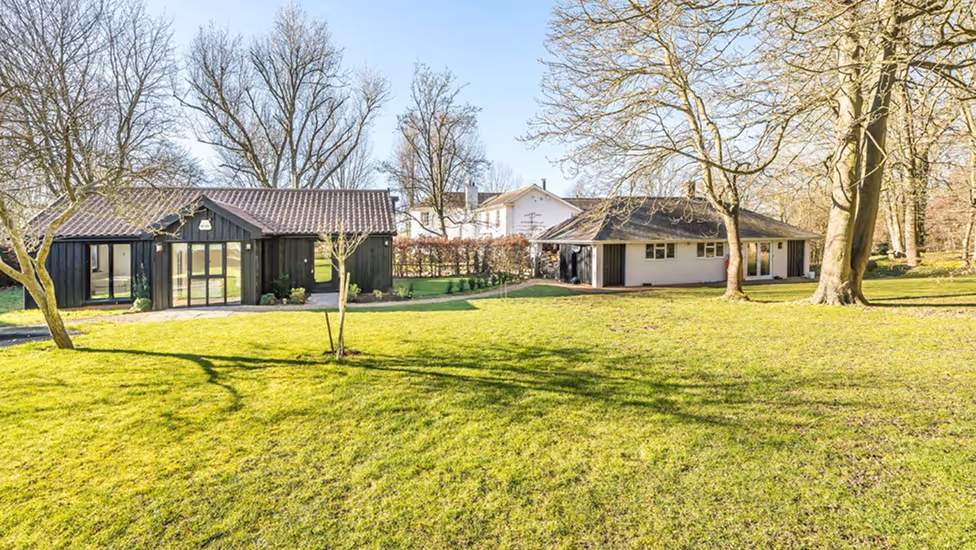 Two modern single-story houses with dark wood and white facades set in a spacious yard with green grass and leafless trees under a clear blue sky.