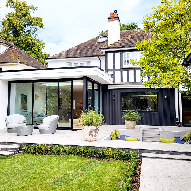 Modern house with large glass sliding doors, outdoor seating area, and a small sunken patio surrounded by greenery.