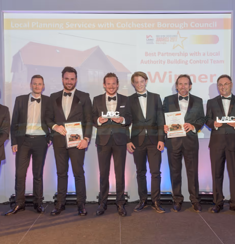 Group of men in tuxedos standing on stage holding awards in front of a screen showing 'Best Partnership with a Local Authority Building Control Team' winner announcement.
