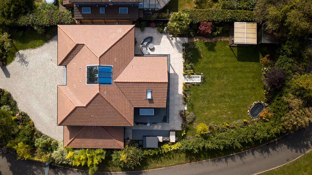 Aerial view of a modern house with a brown tiled roof, patio furniture, a driveway, and a green garden enclosed by shrubs and trees.