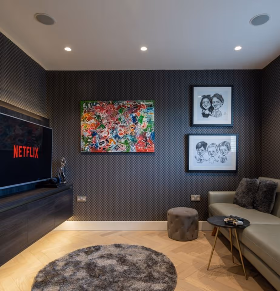 Modern living room with dark textured walls, a large TV displaying Netflix, colorful abstract and black-and-white caricature artwork, gray round rug, beige sofa with pillows, small round tables, and a gray ottoman.