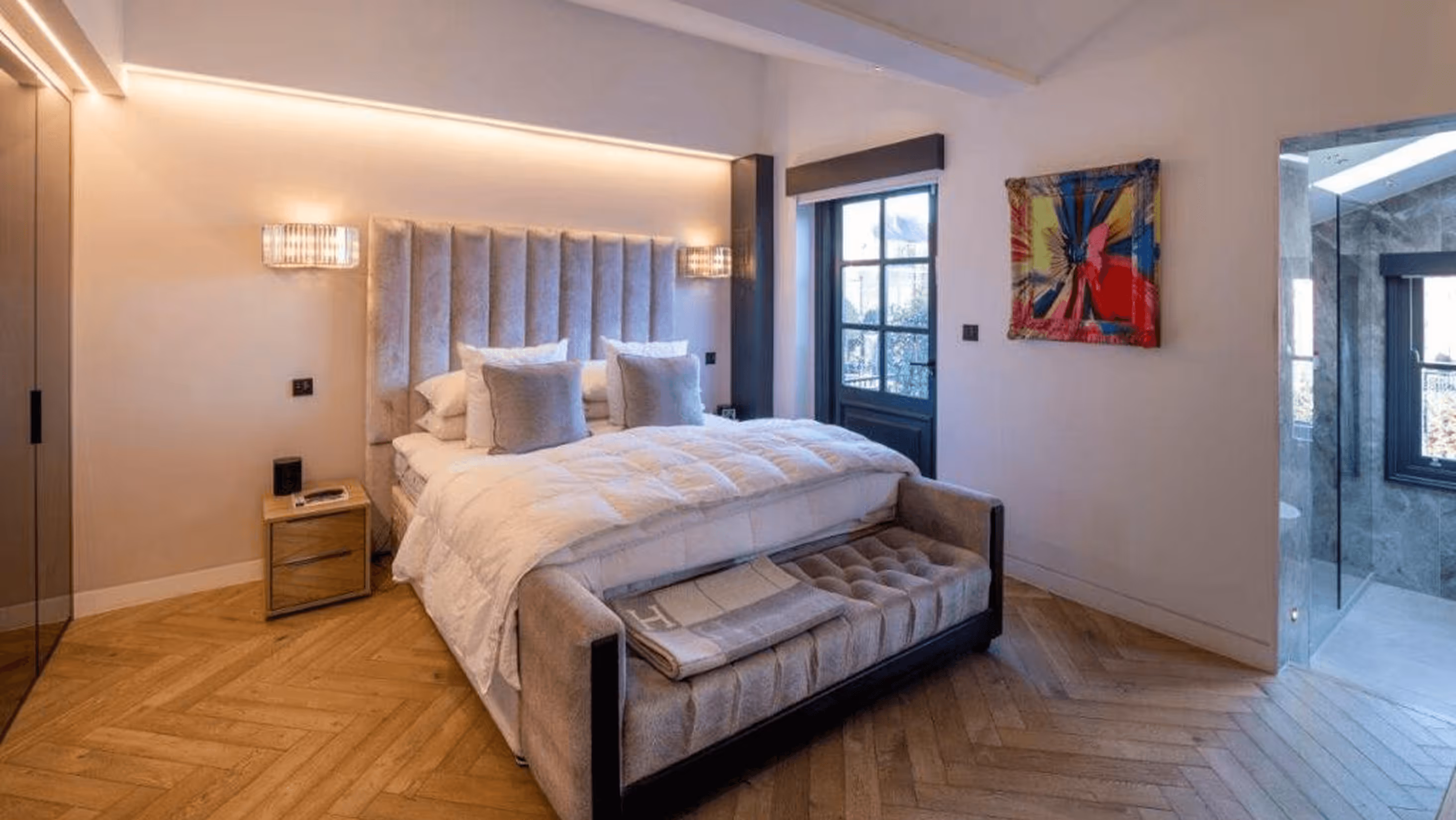 Modern bedroom with padded headboard bed, wooden parquet floor, nightstand, abstract wall art, and attached glass bathroom.