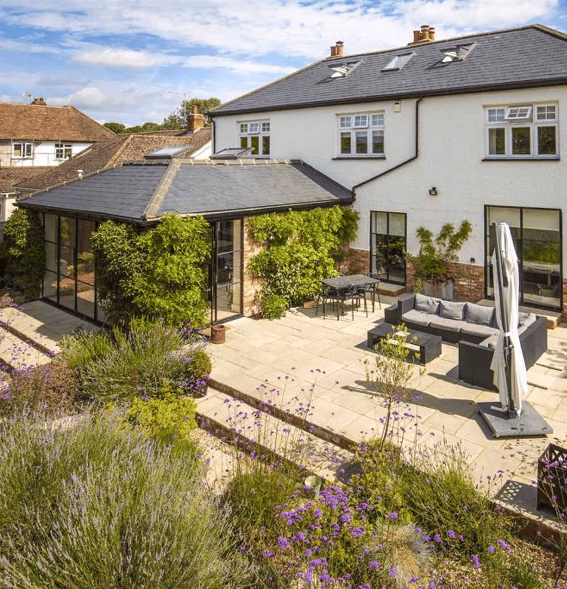 Modern patio with outdoor seating, table, umbrella, and abundant greenery in front of a two-story house with white walls and slate roof.