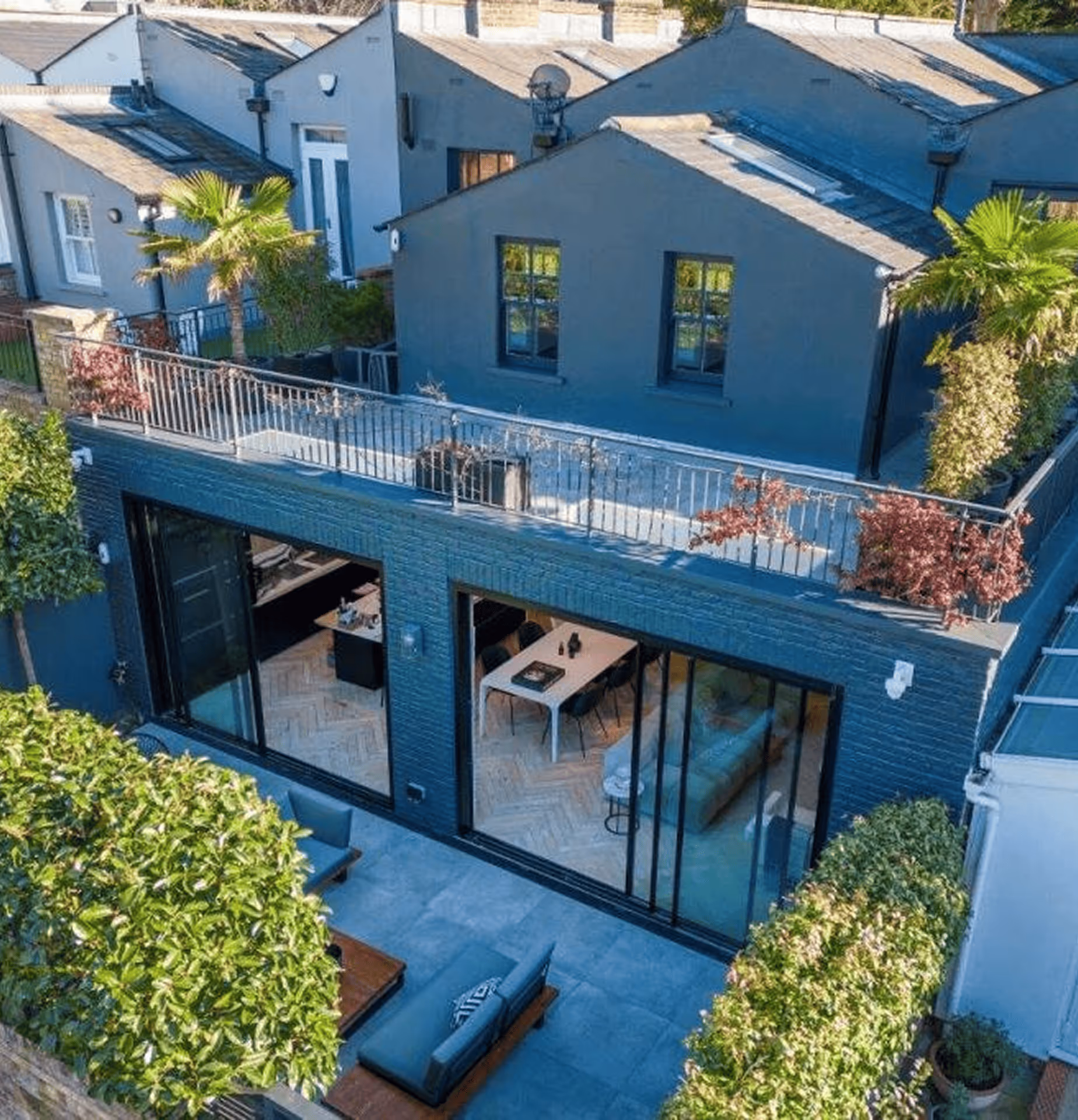 Modern gray house with large glass sliding doors opening to a patio with outdoor seating and green hedges.
