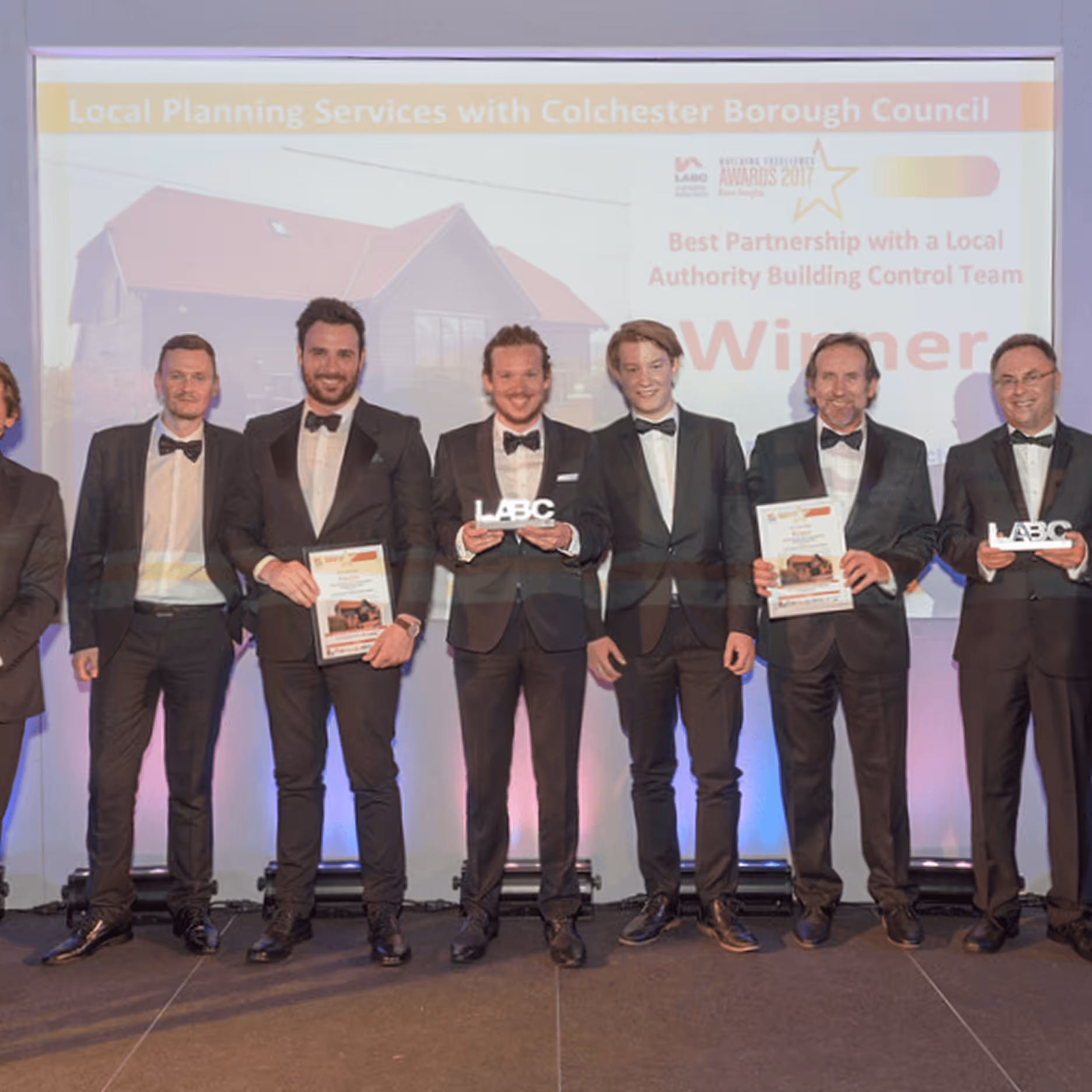Group of men in tuxedos standing on stage holding awards with a backdrop showing a house and text about a local planning services award.
