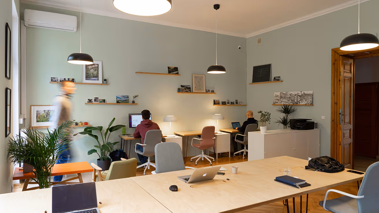 Modern co-working space with people working on laptops and a person walking past indoor plants and shelves with framed pictures.