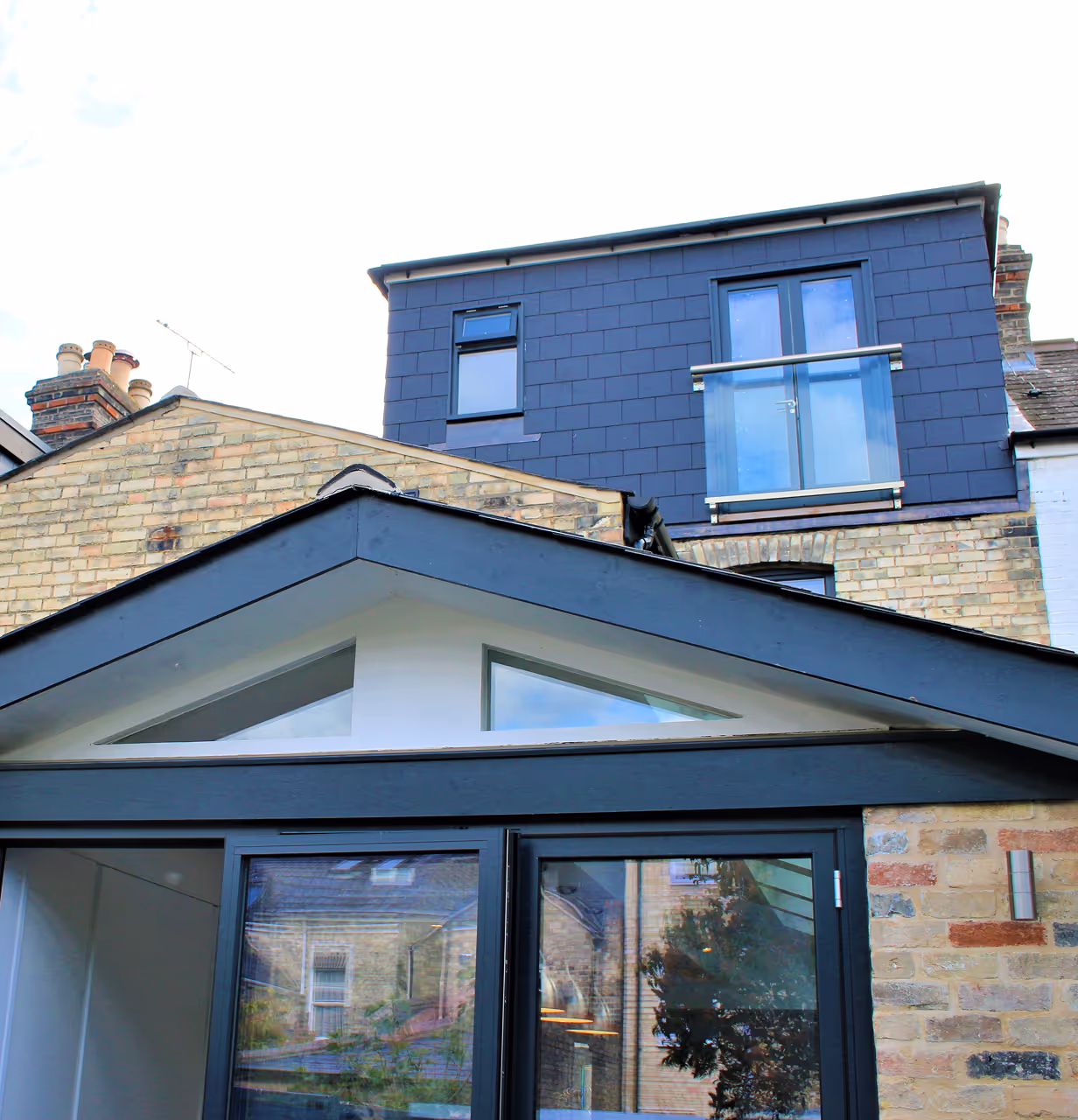 Modern house extension with black framed glass doors and triangular transom windows, attached to a brick building with a dark upper floor featuring a balcony.