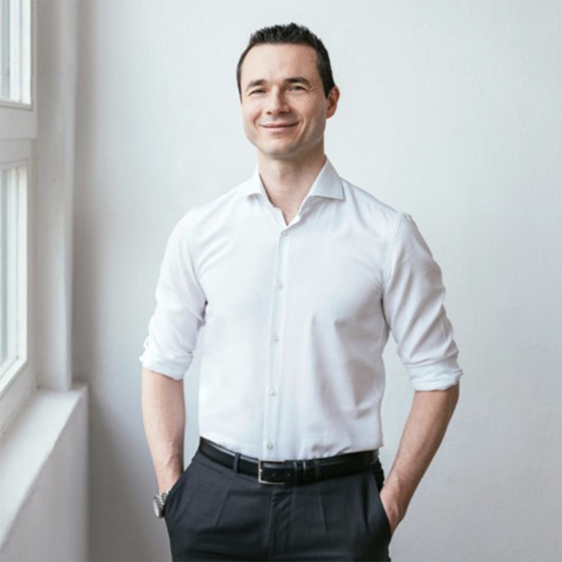 Smiling man standing with hands in pockets wearing a white shirt and black pants by a window.