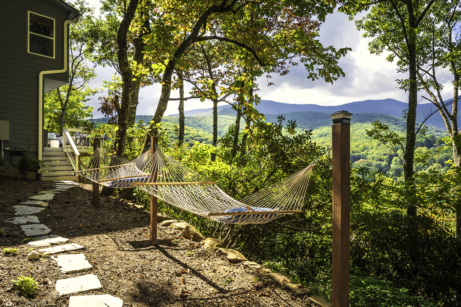 Two hammocks set between wooden posts beside a house overlooking lush green mountains under partly cloudy sky.