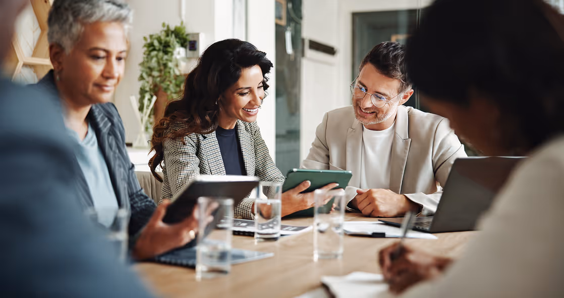 Four diverse professionals sitting around a table in a modern office, two smiling and discussing using tablets.