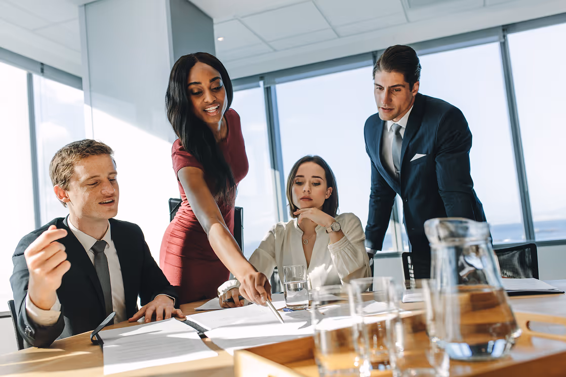Four business professionals in a meeting room reviewing documents at a table near large windows.