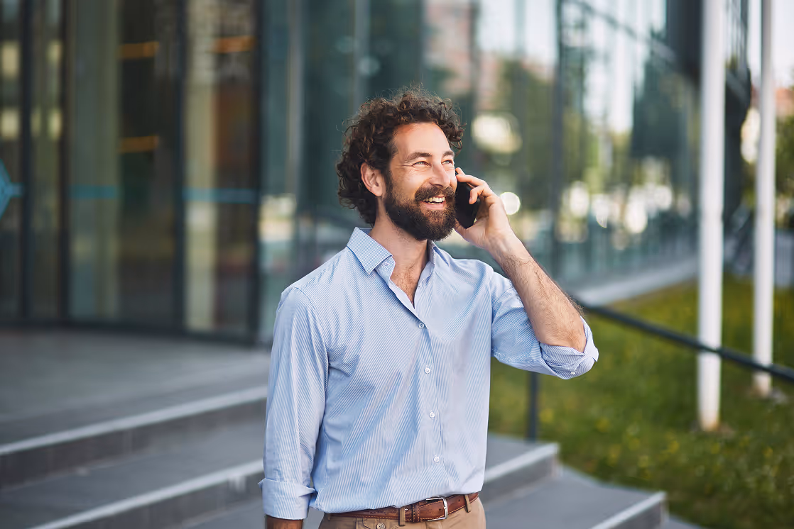 Smiling bearded man in a light blue shirt talking on a smartphone outdoors near steps and glass building.