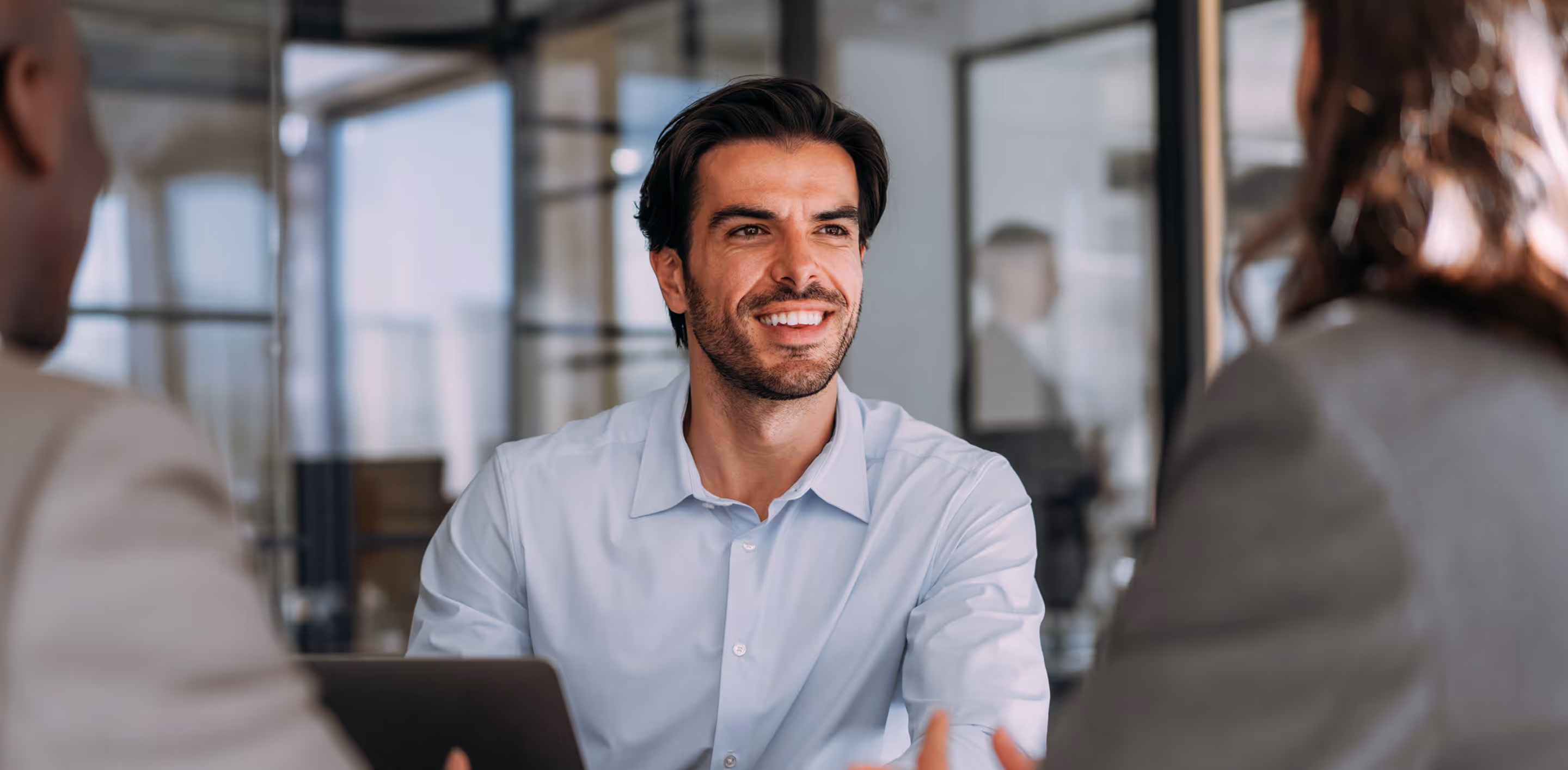 Smiling man in a light blue shirt engaging in a conversation with two colleagues in an office.