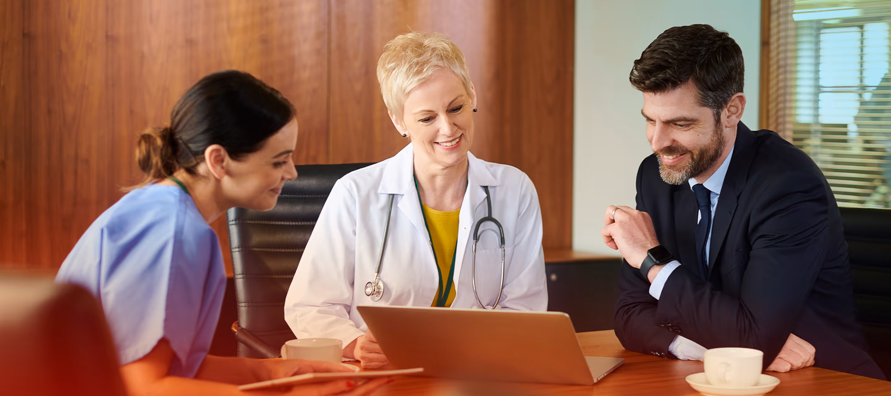 Two female healthcare professionals and a man in a suit sitting at a table looking at a laptop and smiling.