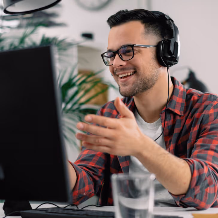 Smiling man wearing glasses and headphones sitting at a desk, engaging with a computer screen.